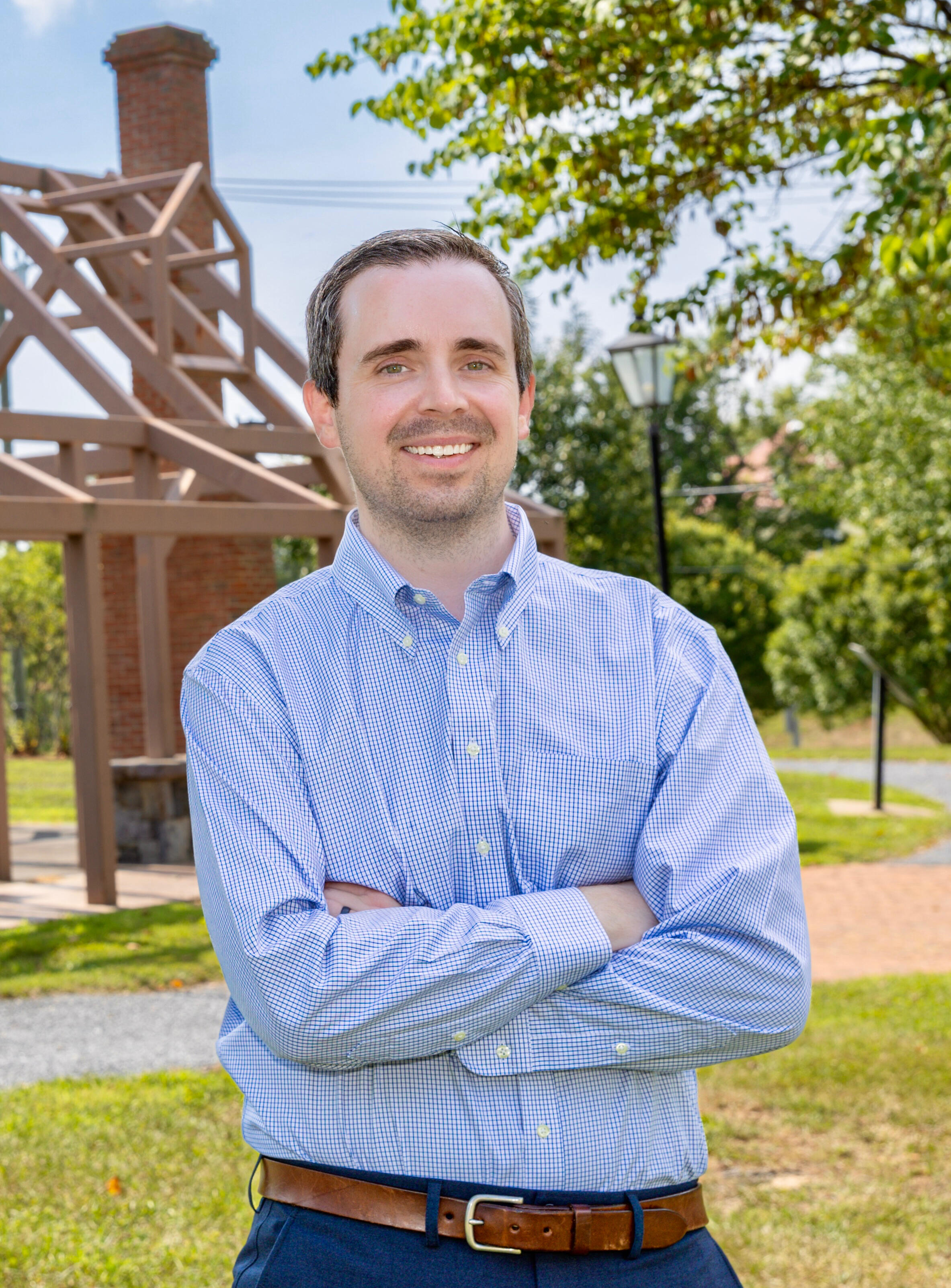 Ethan Wechtaluk standing outdoors in front of a wooden and brick pavilion structure, wearing a light blue button-down shirt and navy pants, smiling with hands in pockets on a sunny day.
